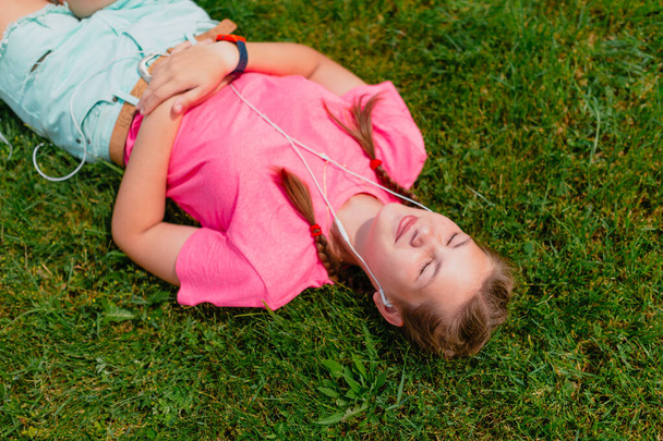 Caucasian girl in a pink T-shirt and white headphones listens to music. Girl student lies smiling on the green grass, turning her face to the sun's rays. - Fotoğraf, Görsel