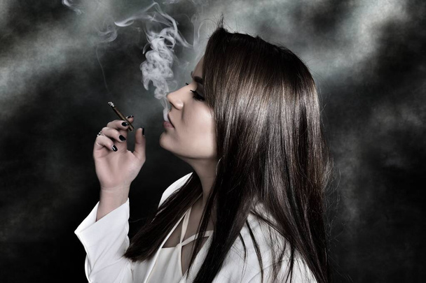 Attractive brunette girl posing in studio and smoking a cigar. Noir, retro style photography. Glamour, lifestyle, fashion concept. - 写真・画像