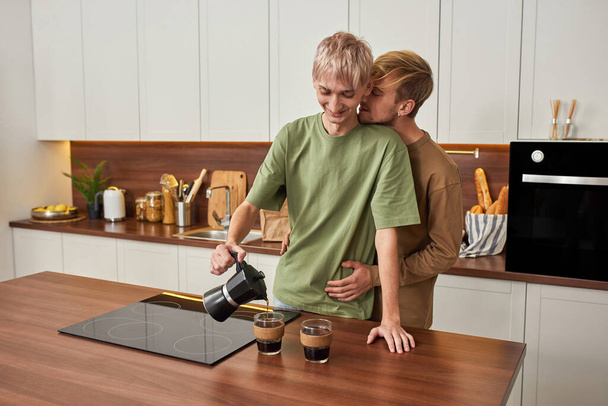 Tierna pareja homosexual masculina abrazándose mientras beben café de la mañana en la cocina moderna en el interior - Foto, Imagen