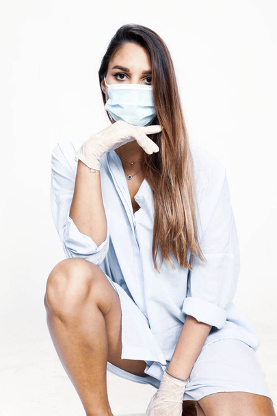 Beautiful brunette female model posing in studio and wearing a protective medical mask and gloves due to Corona virus on isolated white background. - 写真・画像