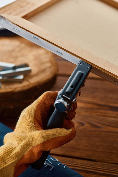 Мужская рука в перчатке держит степлер, крупный план, деревянный фон. Профессиональные инструменты, столярное оборудование, деревообрабатывающие инструменты - Фото, изображение
