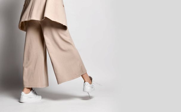 Legs of woman in beige business smart casual suit, sneakers walking over light. Back view. Stylish business female wear - 写真・画像