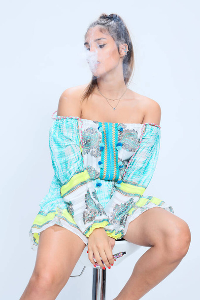 Hermosa morena modelo femenina posando en el estudio y fumando un cigarro eléctrico sobre un fondo aislado. Estilo, fumar, concepto de moda. - Foto, imagen