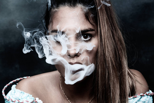 Beautiful brunette female model posing in studio and smoking an electric cigar on isolated background. Style, smoking, fashion concept. - 写真・画像