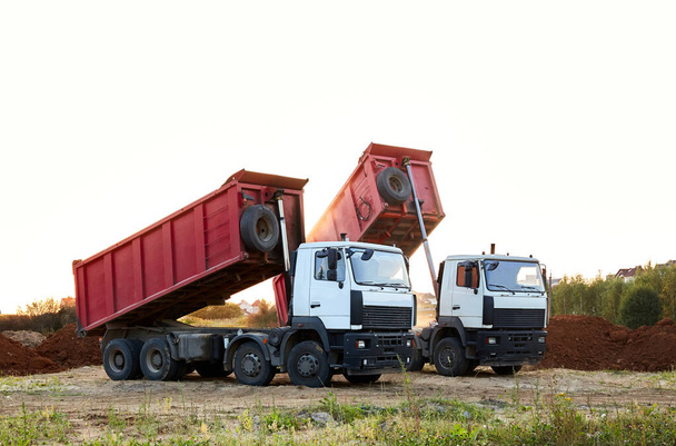 Twee rode vuilniswagens tilden tegelijkertijd de lichamen op om het zand te lossen. Vrachtvervoer. Grote multi-ton truck. Vracht lossen. Bouwplaats en machines. Een spandoek. Gemeenschappelijk standpunt. - Foto, afbeelding