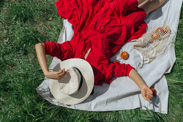 Beautiful girl in red dress lying on green grass, summer picnic with apples, top view - Fotó, kép