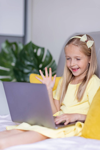 Cute little caucasian girl with blonde hair in fashionable dress illuminating yellow color sitting at home during coronavirus pandemic quarantine and using laptop. Stay at home during covid-19 - Фото, изображение