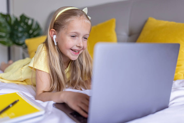 Cute little caucasian girl with blonde hair in fashionable dress illuminating yellow color sitting at home during coronavirus pandemic quarantine and using laptop. Stay at home during covid-19 - Photo, image