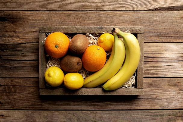 Fresh tropical fruits in a wooden delivery box on a wooden background. Orange, banana, kiwi and lemon top view flat lay with copy space for some advertisement text - Photo, Image