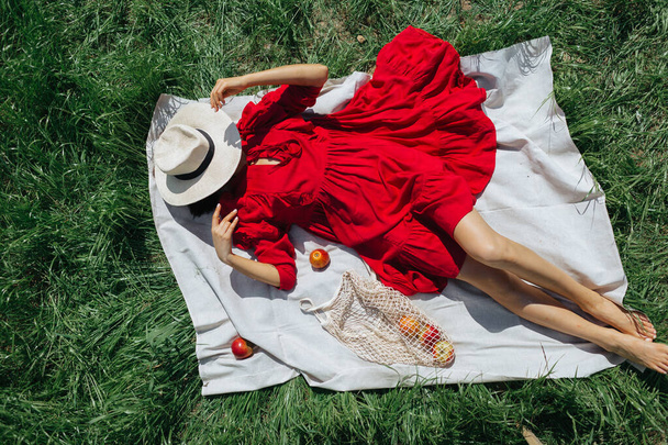 Krásná dívka v červených šatech ležící na zelené trávě, letní piknik s jablky, horní pohled - Fotografie, Obrázek
