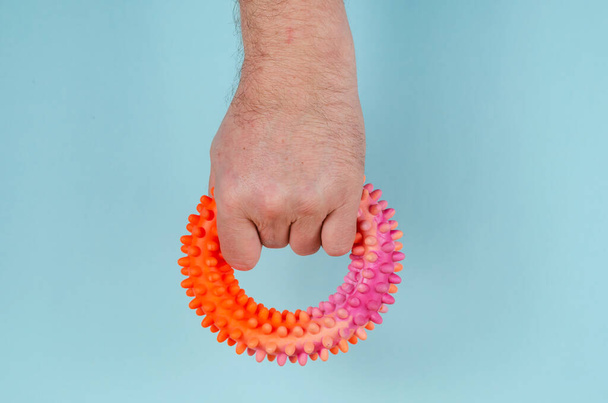 A hand clutching a red rubber expansion band against a blue background. Adult male holding a hand-held round hand trainer. Side view. Sports, active lifestyle. - Photo, Image