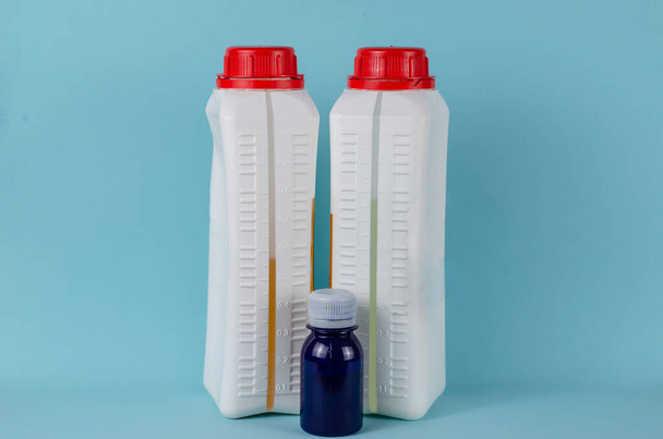 Epoxi líquido en botellas y tinte azul sobre fondo azul. Dos contenedores blancos con tapa. Epoxi y endurecedor. Escala de llenado. Vista lateral. - Foto, imagen