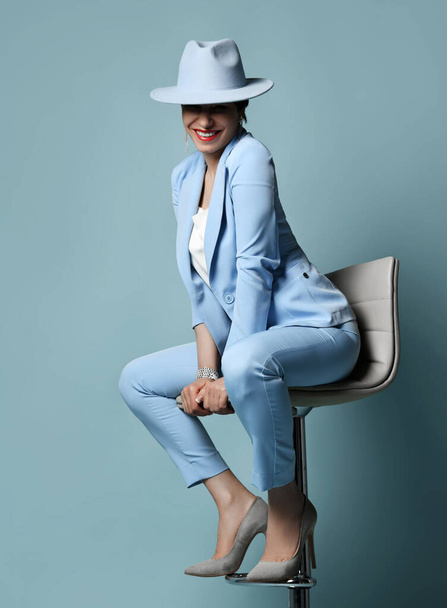 Cheerful brunette woman in blue business suit, covering eyes hat and high-heeled shoes sits on bar stool. Full-growth - Photo, image