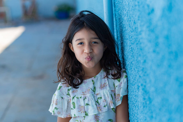 Una chica, una niña, brasileña haciendo pucheros en los labios, con el pelo negro liso, apoyada contra una pared azul. - Foto, Imagen