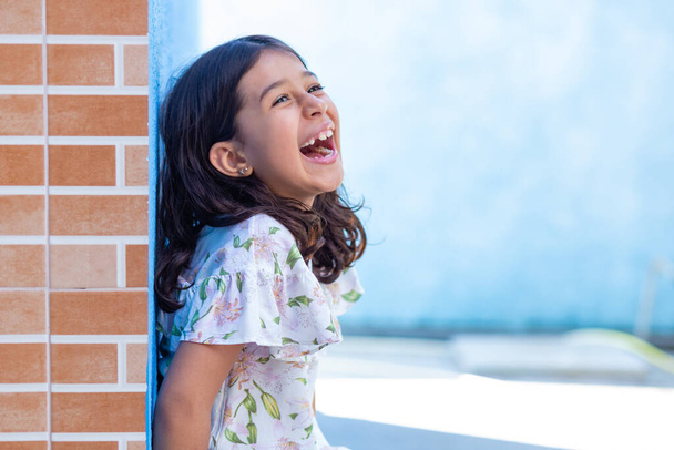 Una chica brasileña sonriente con el pelo negro liso, apoyada contra una pared azul. - Foto, Imagen
