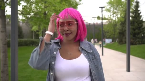 Młoda stylowa czarna kobieta z różowymi włosami spaceruje po parku w słoneczny dzień - Materiał filmowy, wideo