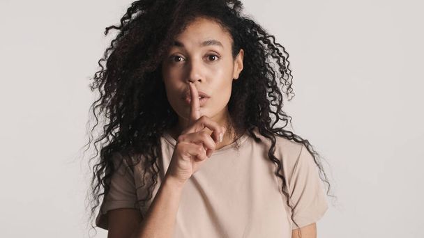 アフリカ系アメリカ人の若い女性が唇の上に指を前に置き、白い背景の上に秘密の表現であることを示す。言うなよ - 写真・画像