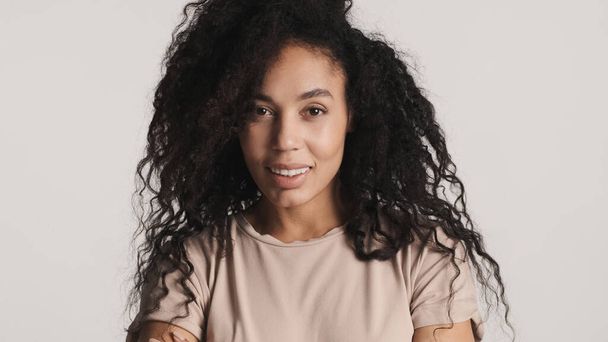Junge wunderschöne Afro-Frau mit dunklen, flauschigen Haaren sieht charmant vor der Kamera isoliert auf weißem Hintergrund aus - Foto, Bild