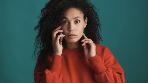 Young serious African American woman dressed in red sweater talking on phone intently looking in camera over blue background - Photo, Image