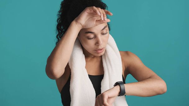 Attractive Afro sporty woman keeping towel on shoulders checking her training result on smartwatch isolated on blue background. Sporty girl - Photo, image