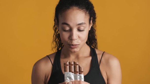 Close up donna afro-americana cercando carino mangiare cioccolato bar dopo l'allenamento su sfondo giallo - Foto, immagini