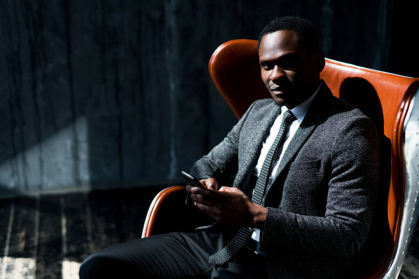 Smiling african american businessman sitting on chair and working on phone while looking at camera - Valokuva, kuva