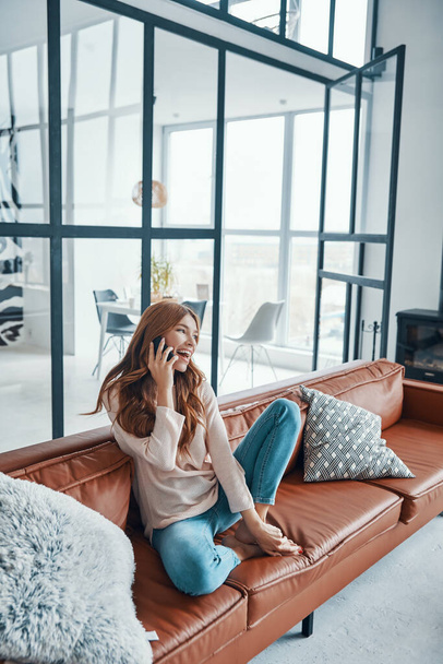 Smiling young woman taking on the phone while sitting on the sofa at home - Foto, Bild