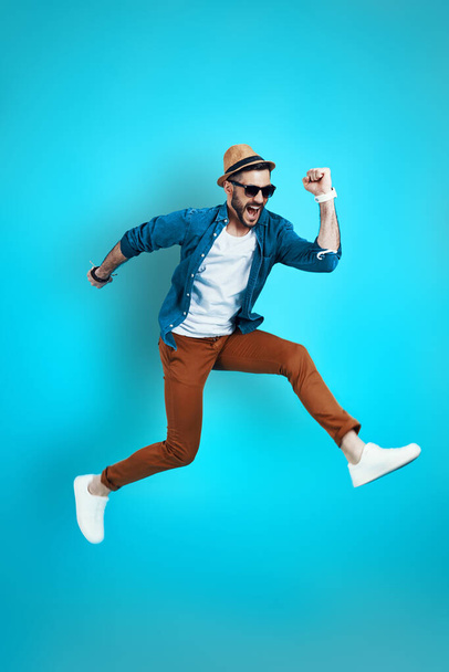 Full length of cheerful young man in casual wear shouting and gesturing while hovering against blue background - Foto, imagen