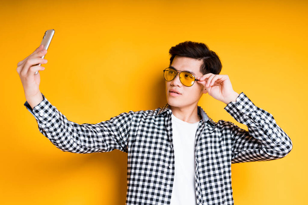 Handsome young asian man wearing bright glasses taking selfie while holding phone in hand - Photo, image