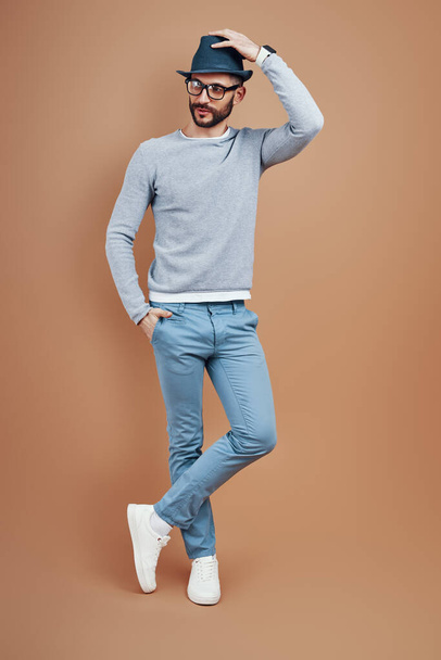 Full length of fashionable young man in casual wear adjusting hat while standing against brown background - Foto, imagen