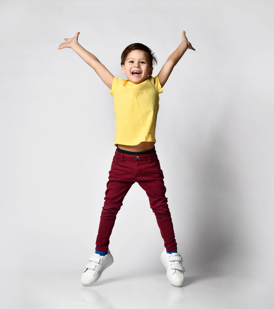 portrait of fashionable preschool little boy in yellow t-shirt, red denim pants, white sneakers, standing isolated on light background - Photo, Image