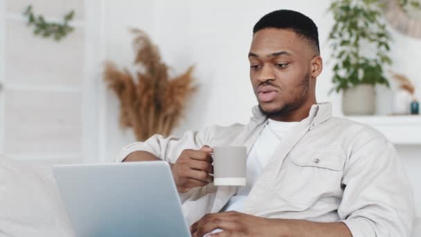 African man young afro student ethnic black male sitting at home holding cup with hot tea coffee looking at laptop screen reads out loud speaks by video chat learns words text, quarantine e-learning - Footage, Video