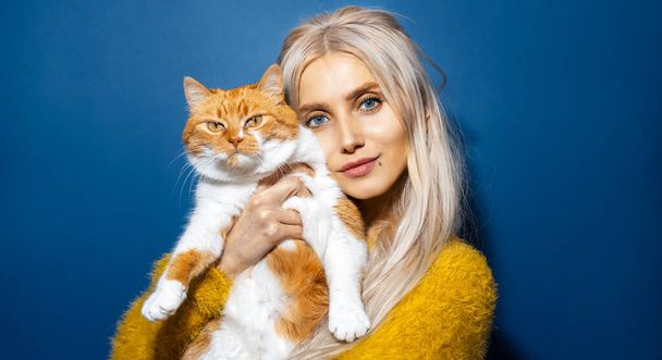Studio ritratto di giovane ragazza bionda che abbraccia il suo gatto bianco rosso su sfondo di colore blu. - Foto, immagini