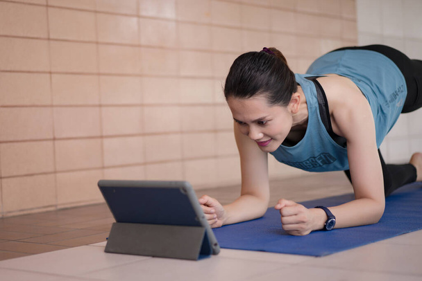 Μια μεσήλικη Ασιάτισσα με χαλαρά αθλητικά ρούχα που κάνει ένα πρόγραμμα προπόνησης άσκησης ακολουθεί ένα online μάθημα προπόνησης σε ένα tablet στο σπίτι κατά τη διάρκεια της πανδημίας COVID-19 και το κλείδωμα της πόλης - Φωτογραφία, εικόνα