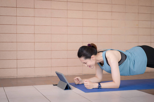 Una mujer asiática de mediana edad en trajes deportivos relajados haciendo un programa de entrenamiento de ejercicio sigue una lección de entrenamiento en línea en una tableta en casa durante la pandemia de COVID-19 y el bloqueo de la ciudad - Foto, Imagen