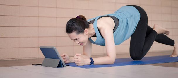 A middle-aged Asian woman in relaxed sports outfits doing an exercise workout training program follows an online workout lesson on a tablet at home during the COVID-19 pandemic and city lockdown - Photo, Image