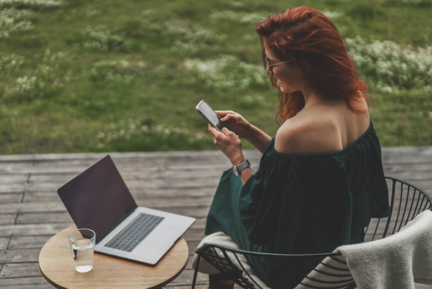Вид сбоку бизнес-леди со смартфоном и ноутбуком, работающей на террасе  - Фото, изображение