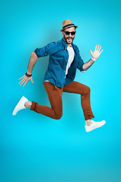 Full length of playful young man smiling and gesturing while hovering against blue background - Фото, изображение