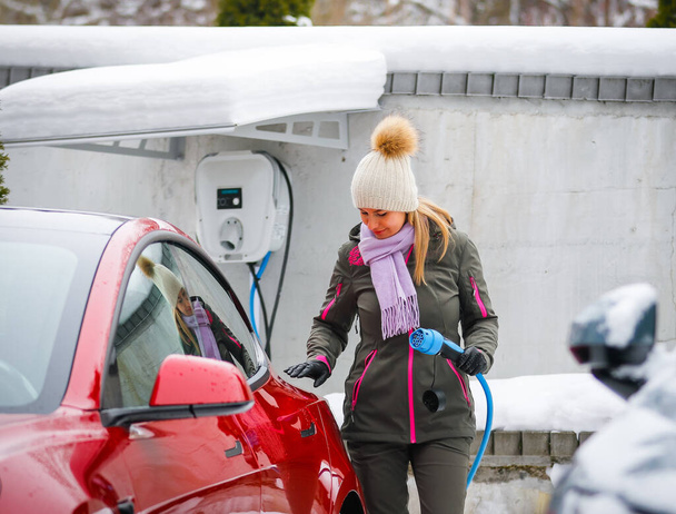 Stylish girl stands near her red electric car and waits for the car to charge. Connecting the plug of the electric vehicle charger. - Photo, Image