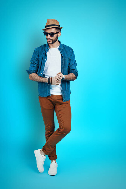 Full length of stylish young man wearing hat and looking away while standing against blue background - Фото, изображение