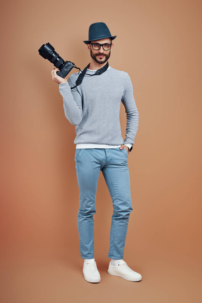 Full length of charming young man in casual wear smiling and looking at camera while standing against brown background - Fotoğraf, Görsel