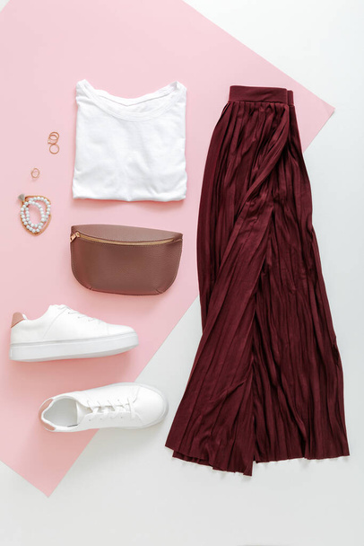 Female spring look summer outfit burgundy skirt white shoes sneakers white basic tshirt waist bag. Folded fly clothes for women fashion urban basic outfit with accessories on pink background. Top view - Foto, afbeelding