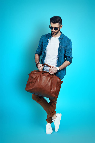 Full length of handsome stylish young man adjusting bag while standing against blue background - Photo, image