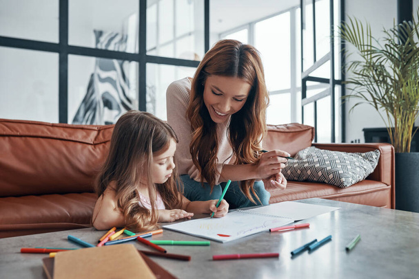Young beautiful mother and her little daughter drawing in sketchbook while sitting on the sofa ay home - Fotoğraf, Görsel