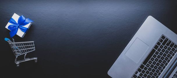 Compras web. Ordenador portátil, carrito de compras y regalo blanco con cinta azul sobre fondo oscuro. Concepto de comercio electrónico y experiencia del cliente. Compra por Internet, tienda online - Foto, imagen