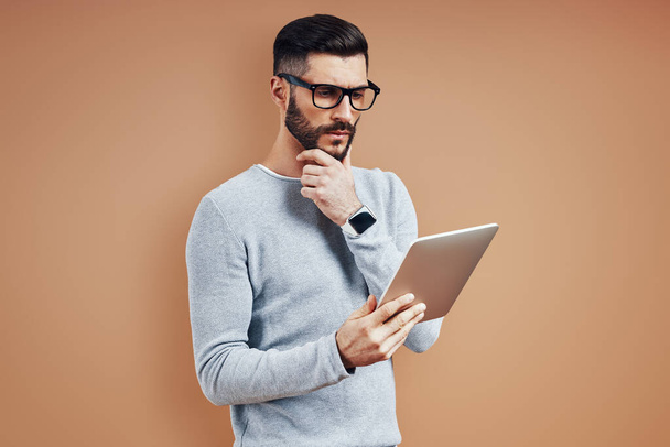 Thoughtful young man in smart casual wear keeping hand on chin and using digital tablet while standing against brown background - 写真・画像