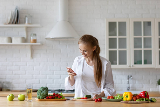 Happy woman chatting online with friends while prepares fresh vegetables stay home, healthy food. - Фото, изображение