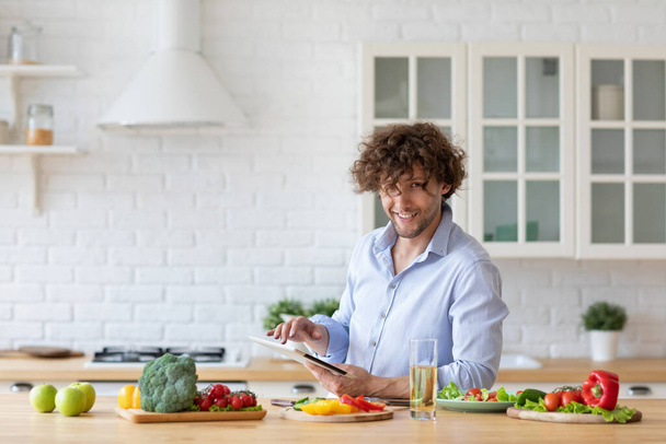Hombre joven viendo video receta en la tableta digital mientras cocina verduras en la cocina. Cuidar de su salud. - Foto, Imagen
