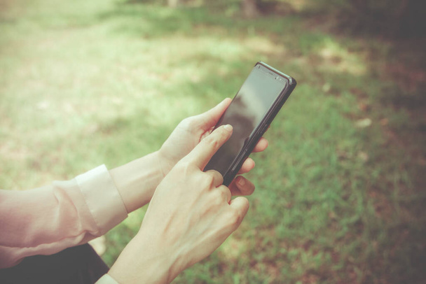 Smartphone Concept Παντού, η Γυναίκα χρησιμοποιεί το χέρι της για να αγγίξει την οθόνη για να αναζητήσει μηνύματα ή άλλες πληροφορίες στην εφαρμογή, ενώ κάθεται στο πράσινο γκαζόν. - Φωτογραφία, εικόνα