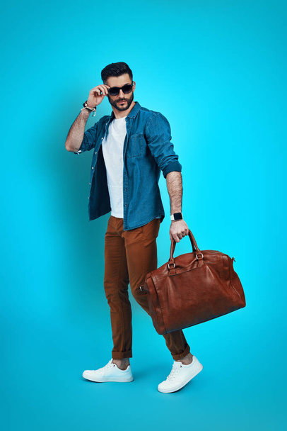 Full length of stylish young man smiling and adjusting eyewear while walking against blue background - Zdjęcie, obraz